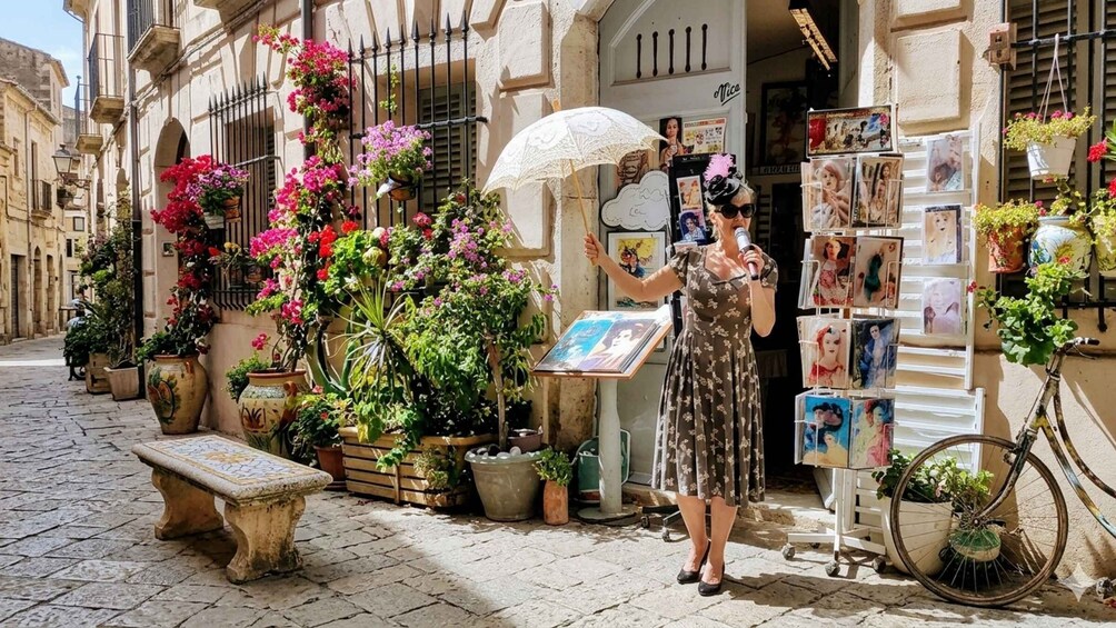 A woman in a floral dress holding a microphone and an umbrella, standing in front of a shop with postcards and plants. 3