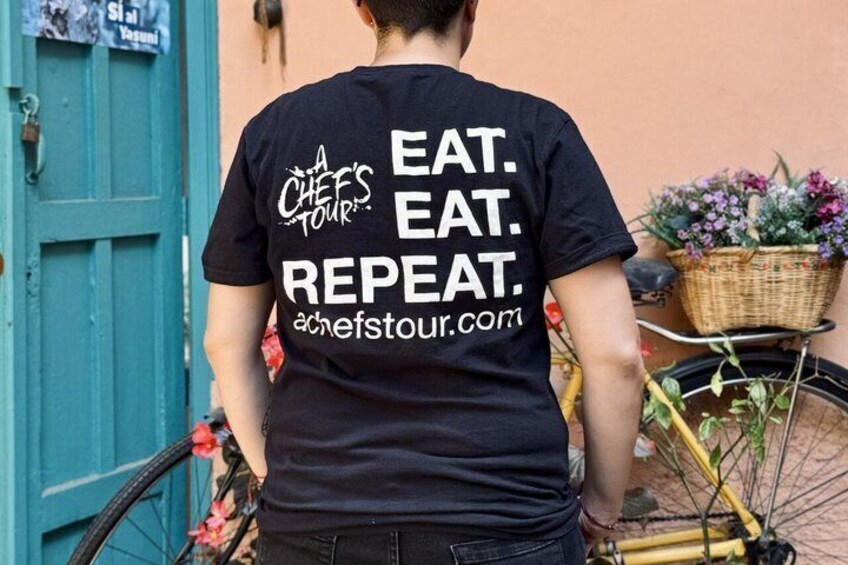 A person wearing a black t-shirt with the text 'A CHEF'S TOUR EAT. EAT. REPEAT.' and a website address. 4