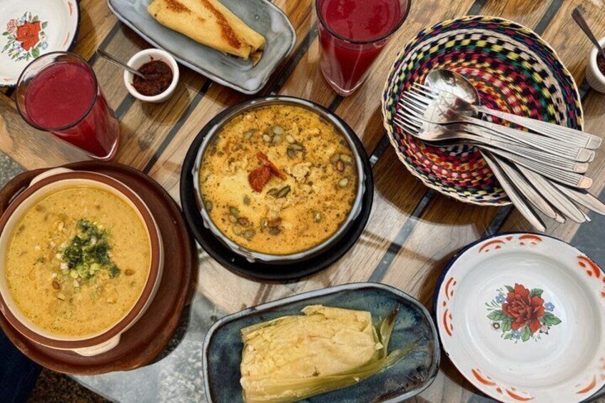 A table with various dishes, including a bowl of soup, a plate of tamales, and a pan of fried food. 1