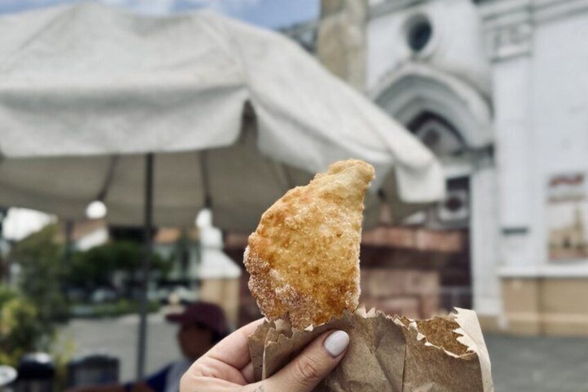 A hand holding a piece of fried food, with a building and an umbrella in the background. 3
