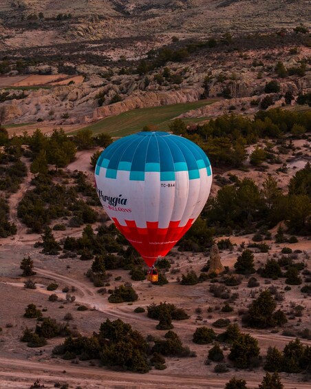 A hot air balloon with 'Chrygia Balloons' branding, flying over a desert landscape with sparse vegetation and rocky terrain. 1