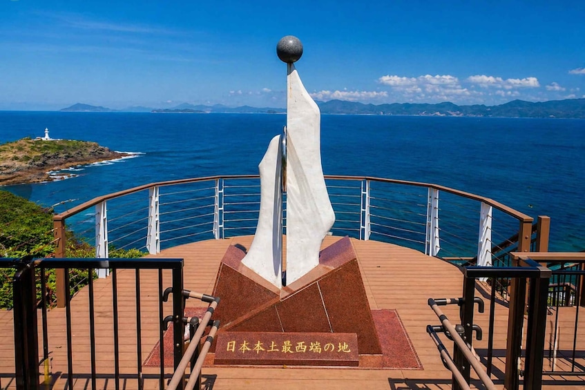 A wooden observation deck with a white flag and a plaque in Japanese, overlooking a coastal landscape with a lighthouse and mountains. 4