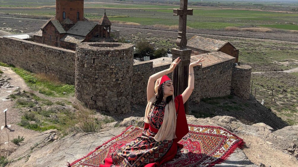A person sitting on a rock with a patterned blanket, a stone church, and a cross in the background. 5