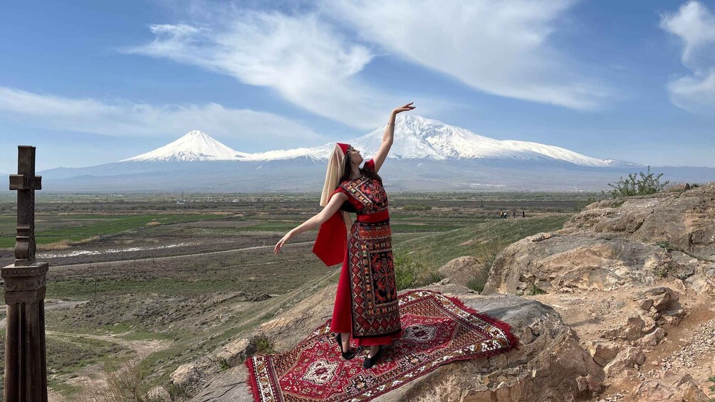 A person standing on a rock with a patterned rug, a cross, and a snow-capped mountain in the background. 3