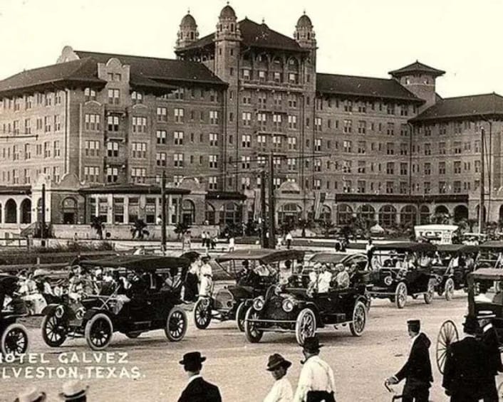 A vintage street scene with early 20th-century cars and a large building with multiple stories and arched windows. 1
