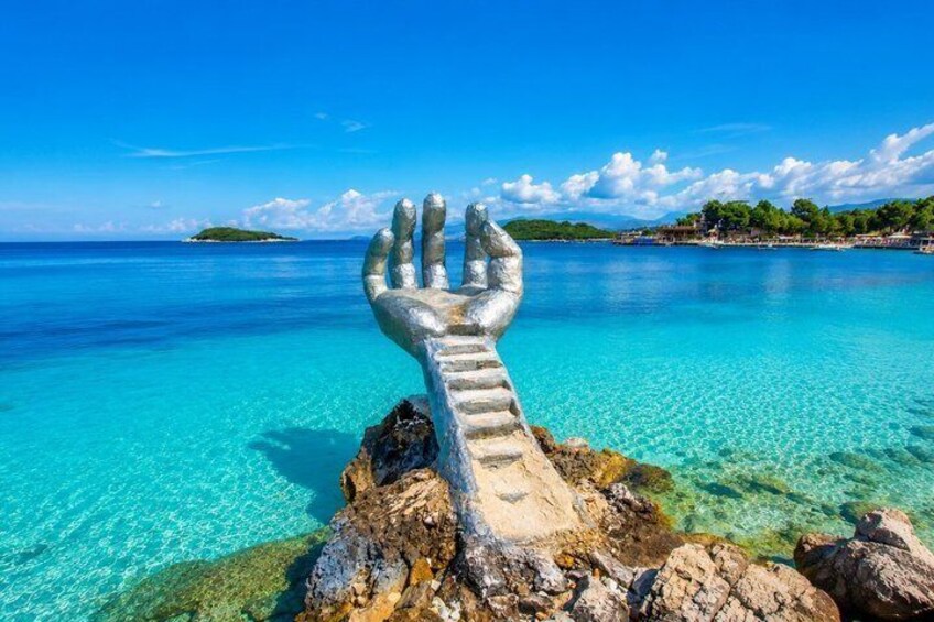 A clear blue sea with a rocky outcrop featuring a large hand sculpture. 1