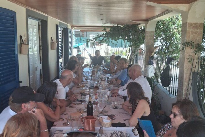 A group of people seated at a long table outdoors, likely at a restaurant or gathering. 1