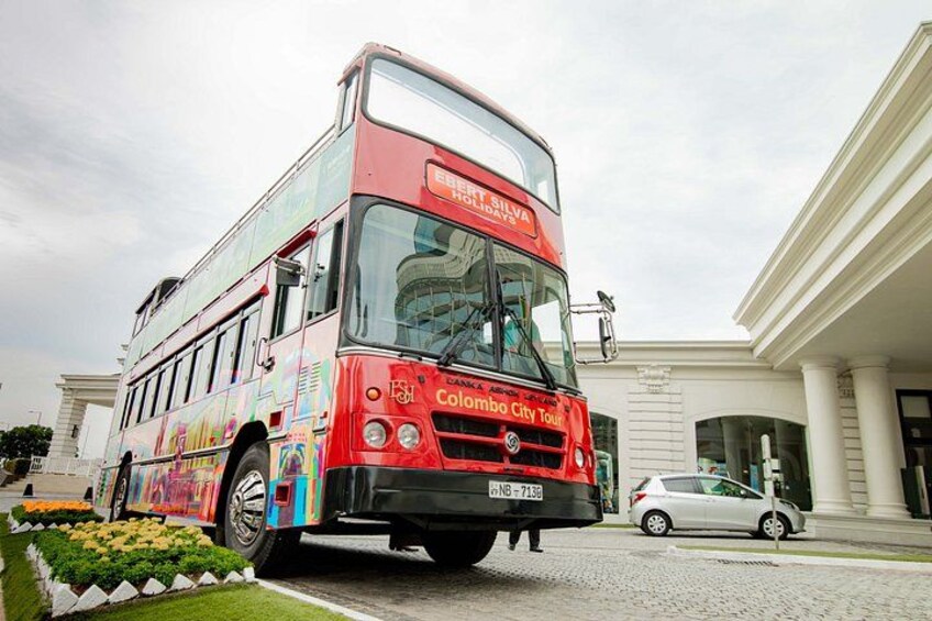 A red double-decker bus with 'Colombo City Tour' written on the front, parked in front of a building with columns. 1