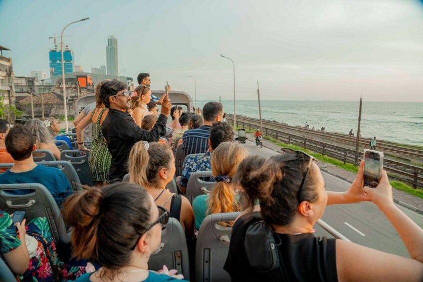 A group of people on a bus, some taking photos, with a coastal road and buildings in the background. 3