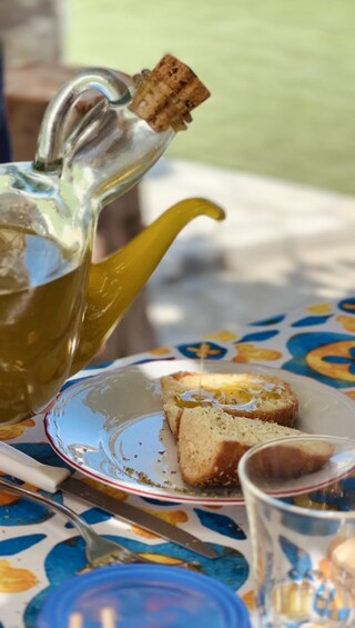 A glass teapot with a cork stopper pouring tea into a cup, a plate with bread, and a fork on a table with a colorful tablecloth. 2