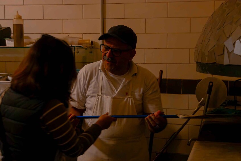 A person in a white chef's uniform holding a blue tool, likely a spatula, in a kitchen with a brick wall and a pizza oven in the background. 2