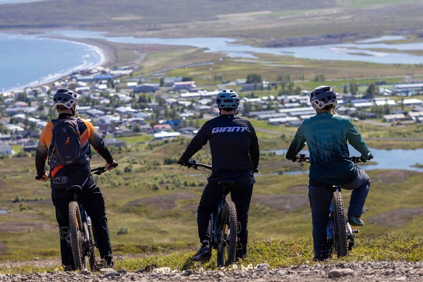 Three mountain bikers on a trail with a view of a coastal town and water bodies in the background. 1