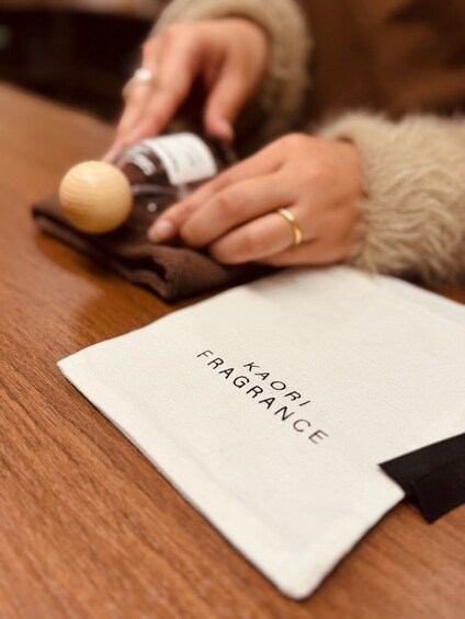 A person holding a small bottle, with a white cloth labeled 'KAORI FRAGRANCE' on a wooden surface. 2