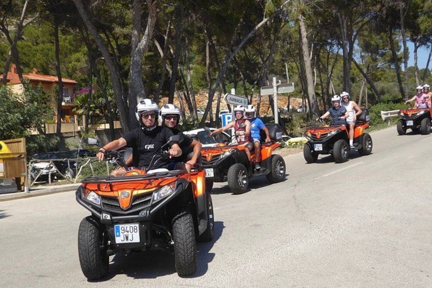 A group of people on quad bikes on a road with trees and buildings in the background. 1