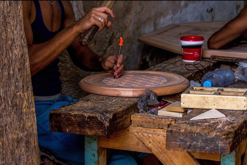 A person carving a wooden object on a circular wooden base with tools and a red container nearby. 5