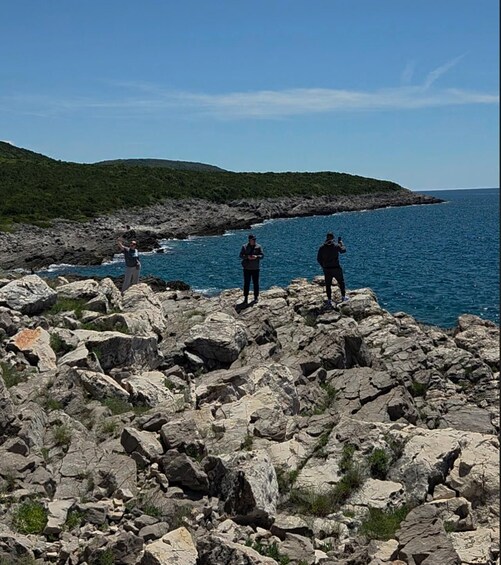 Three people standing on rocky terrain by a body of water with a hilly landscape in the background. 3