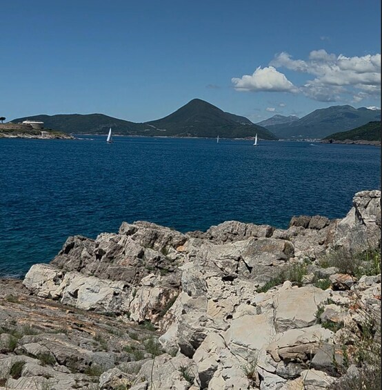A rocky coastline with a clear blue sea, sailboats, and distant mountains. 1