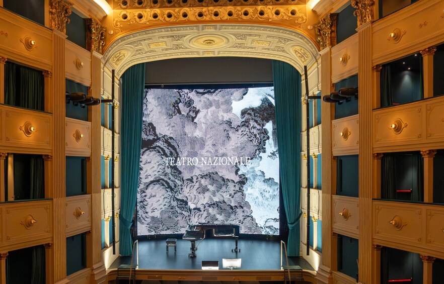 An ornate theater interior with a large screen displaying a map, golden accents, and blue curtains. 1