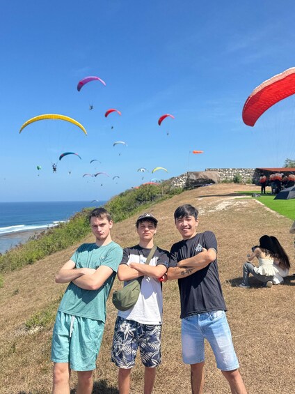 Three people standing on a grassy hill with colorful paragliders in the background. 2