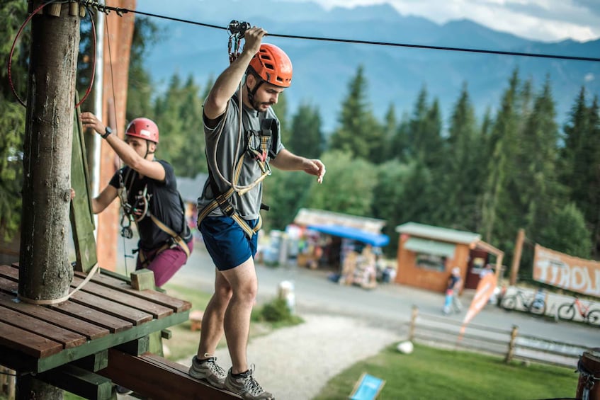 A person rappelling down a wooden platform with safety gear, overlooking a mountainous landscape with a gas station in the background. 5