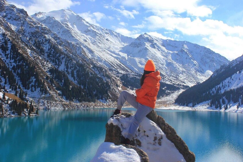 A person in an orange jacket and beanie sitting on a rock by a turquoise lake surrounded by snow-covered mountains. 2