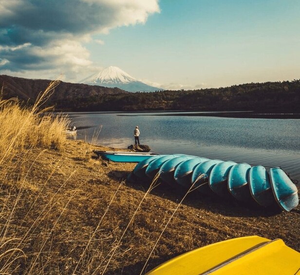 A calm lake with a person standing on a dock, a mountain in the background, and several kayaks and a yellow surfboard on the shore. 2
