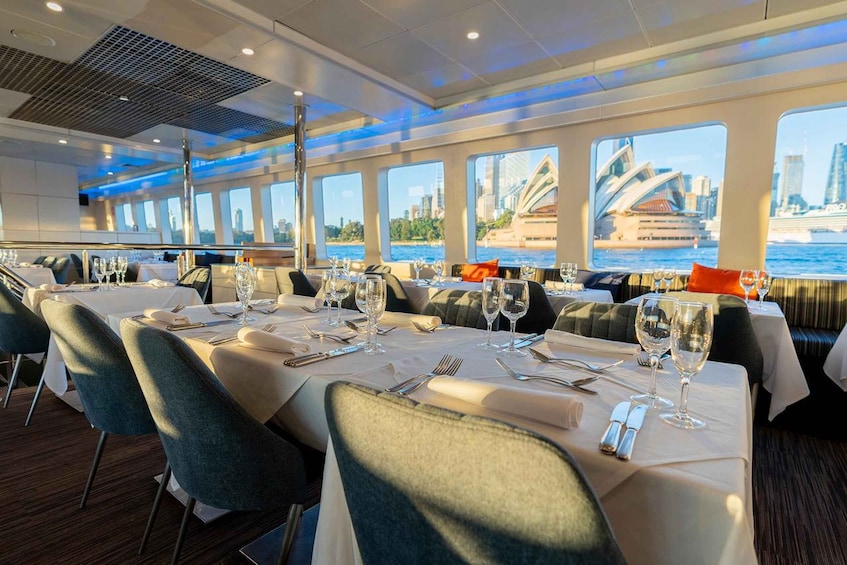 A dining area on a cruise ship with tables set for dinner, overlooking a city skyline with a prominent opera house. 1
