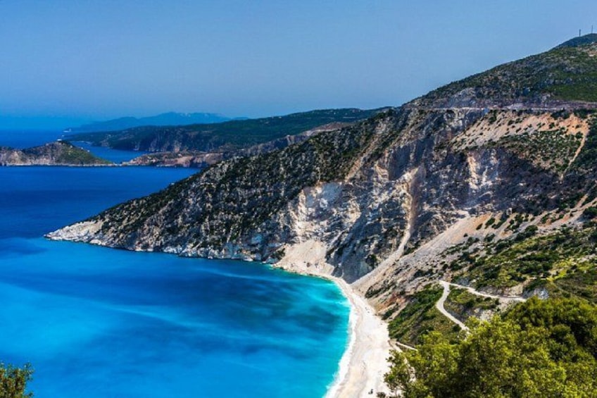 A coastal landscape with a clear blue sea, a sandy beach, and a mountainous terrain with sparse vegetation. 4