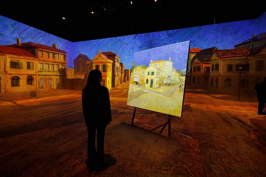 A person standing in front of a painting on an easel in a room with a large projection of a street scene on the wall. 2