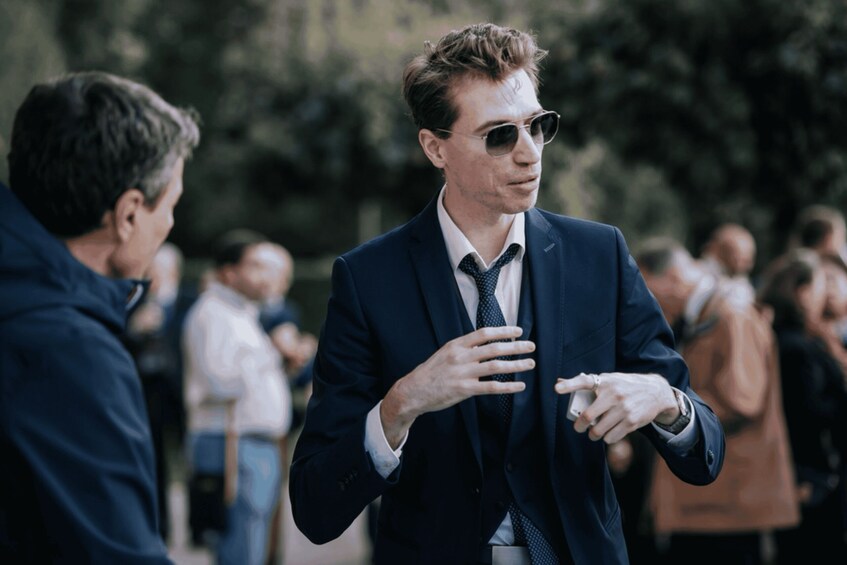 A man in a suit and tie gesturing with his hands, standing outdoors with trees in the background. 3