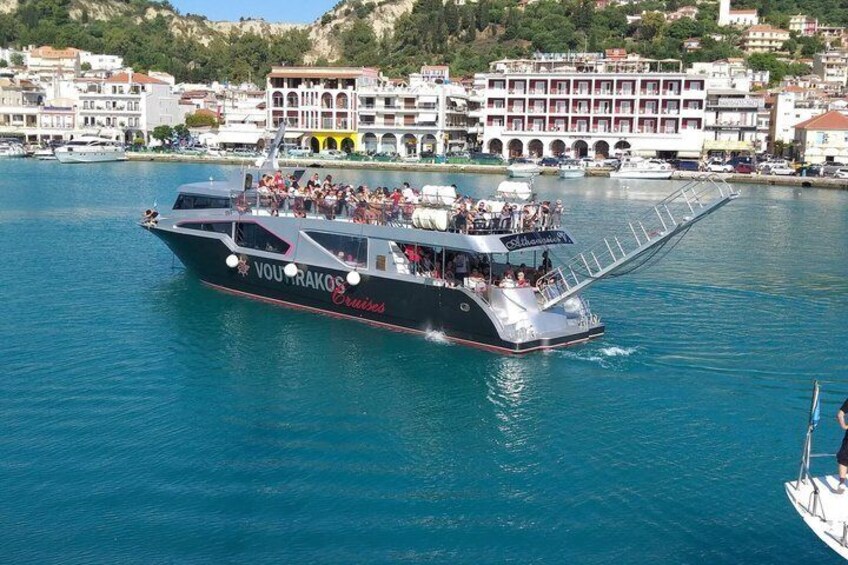 A large boat named 'VOU FRAKOS' cruising on a calm body of water with a cityscape in the background. 2
