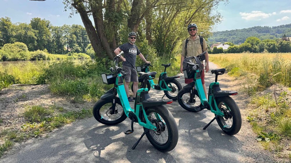 Two people standing next to electric bikes on a gravel path with trees and a field in the background. 4