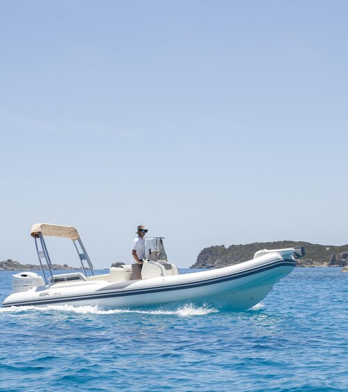 A white motorboat with a canopy on the water, a man steering, and a rocky coastline in the background. 1