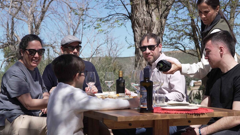 A group of people sitting around a wooden table outdoors, with bottles of wine and glasses on the table. 4