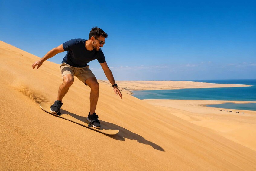 A person snowboarding down a sand dune with a clear blue sky and a body of water in the background. 3