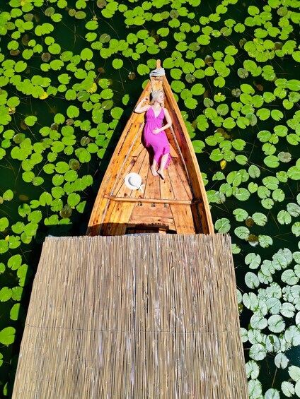 A woman in a pink dress sitting in a wooden boat on a bamboo platform surrounded by lily pads. 4