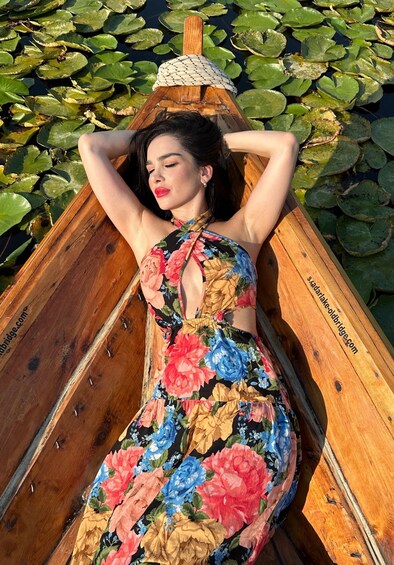 A person in a floral dress lying in a wooden boat on water with lily pads. 2