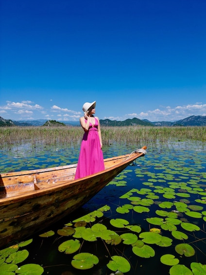 A woman in a pink dress standing in a wooden boat on a lake with lily pads and mountains in the background. 1