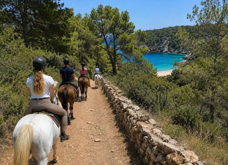 A group of people horseback riding on a dirt path with a stone wall on one side and a beach with clear blue water in the background. 4