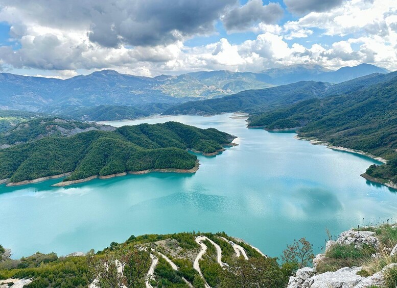 A turquoise lake surrounded by green hills and mountains under a partly cloudy sky. 3