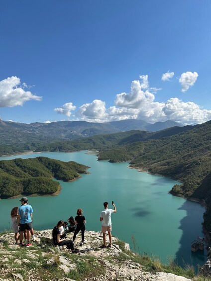 A group of people on a rocky overlook, overlooking a turquoise lake surrounded by green hills. 1