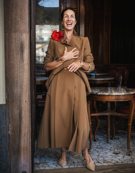 A person in a brown coat with a red flower, standing in a doorway with a tiled floor and wooden furniture. 2