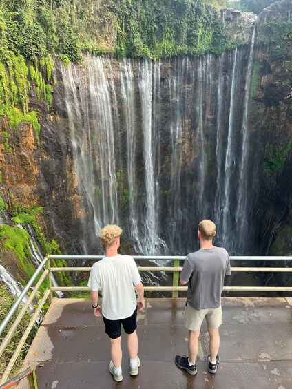 Two people standing on a metal platform, overlooking a multi-tiered waterfall surrounded by lush greenery. 3