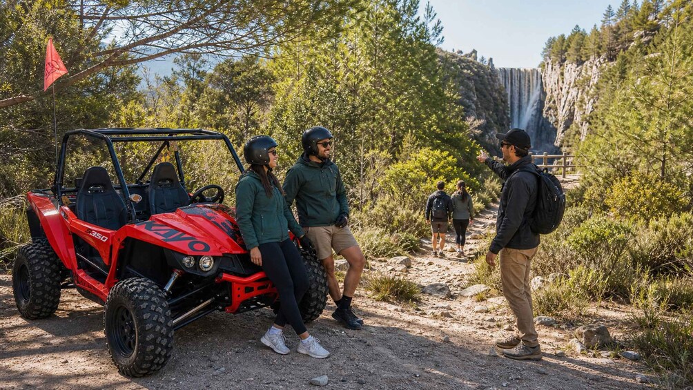 A red off-road vehicle, hikers, and a waterfall in a forested area. 1