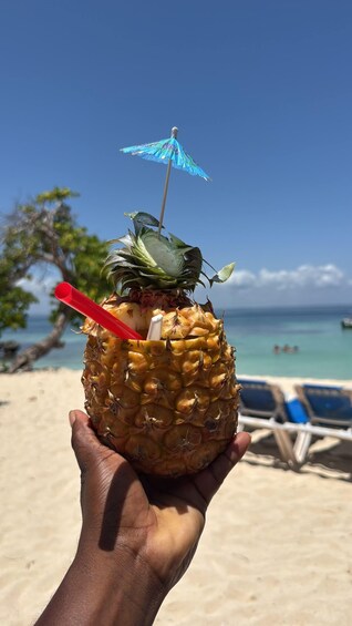 A hand holding a pineapple with a red straw and a blue umbrella on a beach. 3