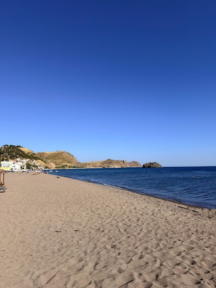 A sandy beach with clear blue water, a few people, and buildings in the background. 4