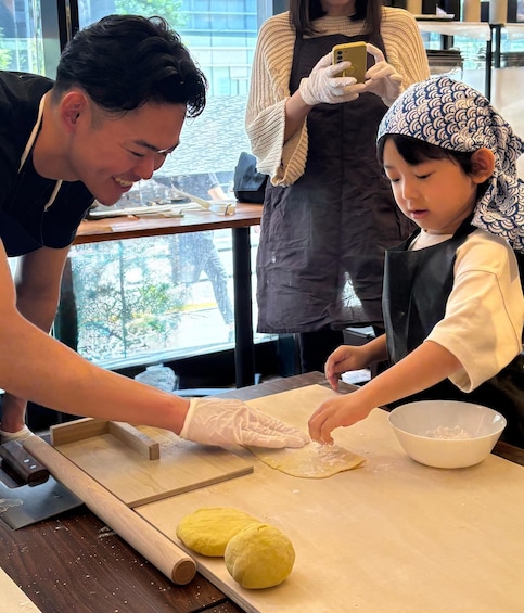 A person teaching a child to roll dough on a table with a rolling pin and a bowl of flour nearby. 1