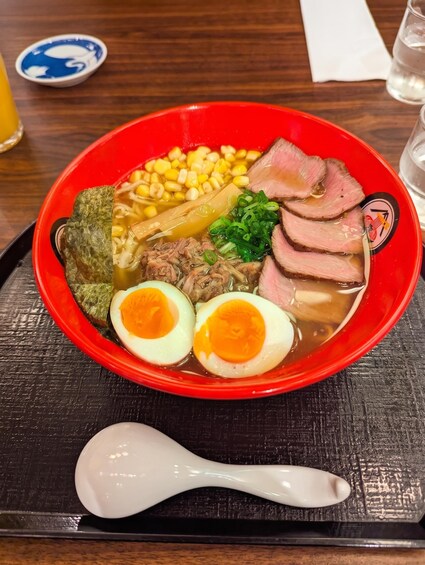 A bowl of ramen with sliced beef, corn, green onions, and a boiled egg on a red plate. 2