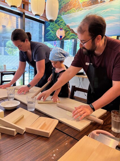 Three people rolling dough on a wooden surface in a kitchen setting. 3
