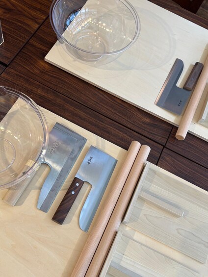 A wooden cutting board with a large cleaver, a smaller cleaver, a rolling pin, and a glass bowl. 2